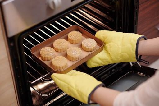 Person wearing yellow oven mitts placing a tray of mooncakes into the oven.