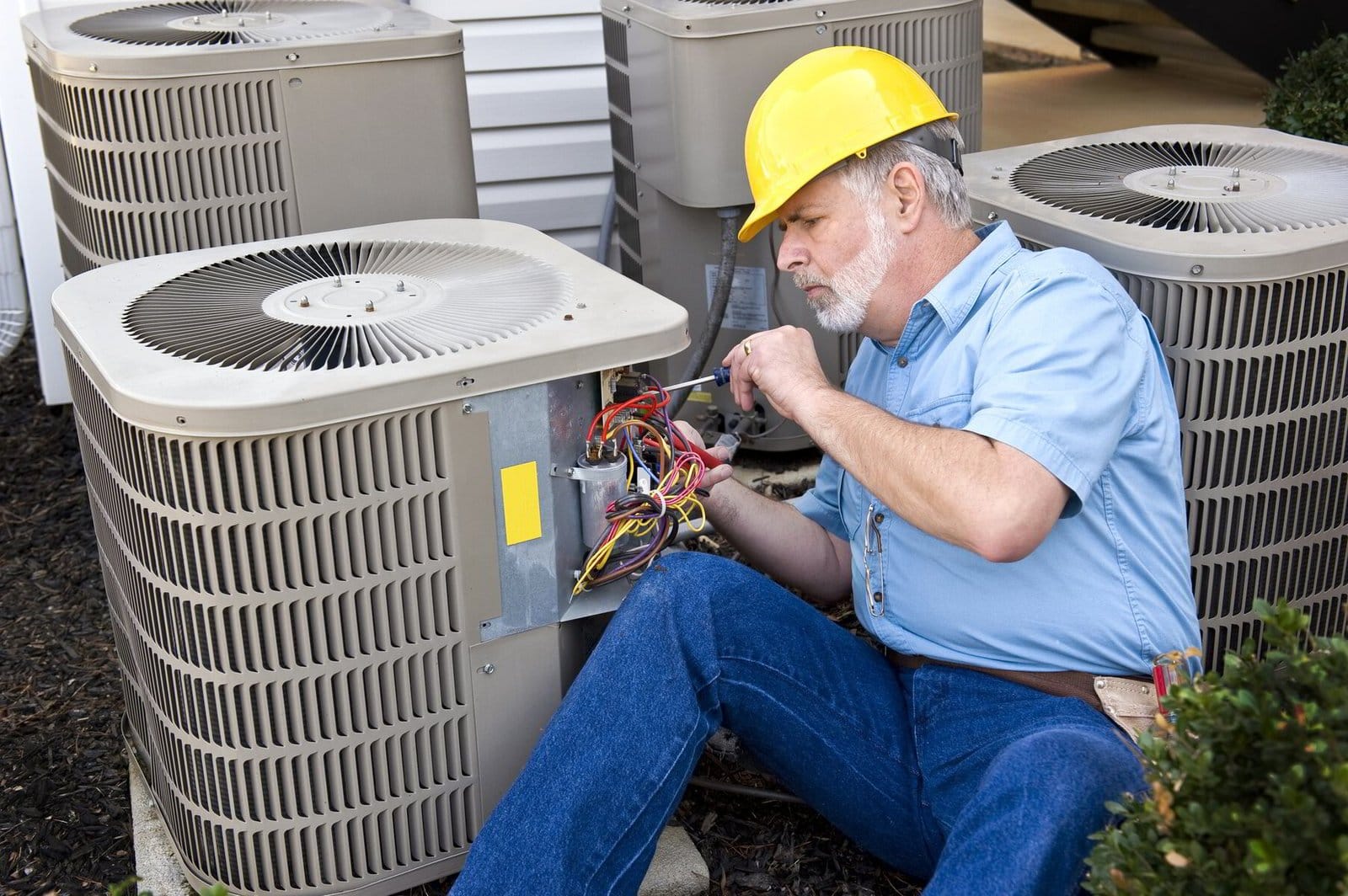 A technician wearing a yellow hard hat is repairing an outdoor air conditioning unit.