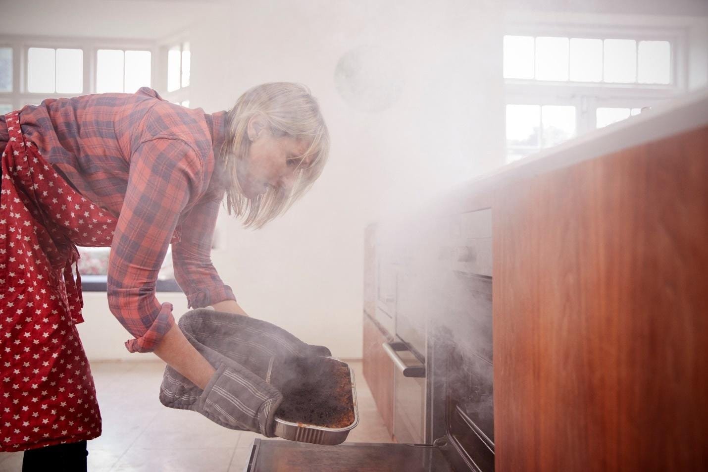 Woman in a red apron removing a smoking dish from an oven, illustrating common causes of a smoking oven and the importance of maintenance.