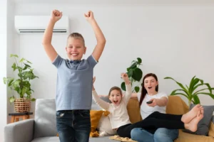 Smiling boy standing with his arms raised in excitement, while a woman and a young girl cheer and watch TV on the couch in a cozy living room with houseplants.