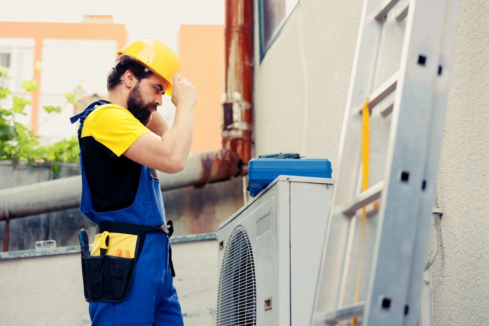 Worker in yellow safety helmet and blue overalls adjusting an air conditioning unit next to a ladder.