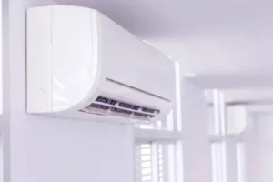 A white air conditioning unit mounted on a wall in a bright room with large windows.
