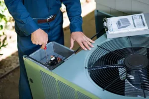 A worker in blue uniform uses a screwdriver to repair an outdoor air conditioning unit, with a manual nearby.