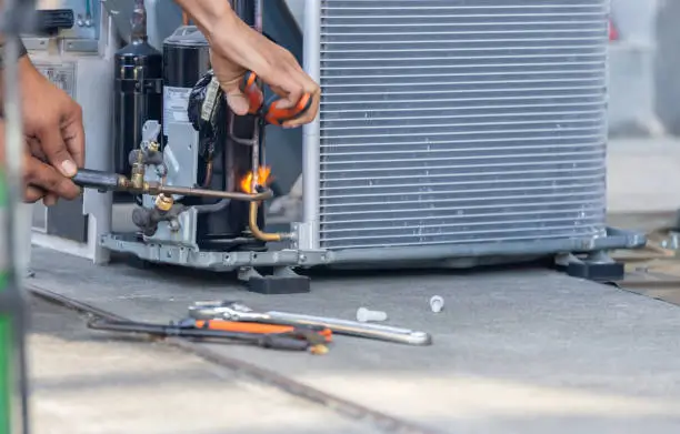 Close-up of a technician using a torch to solder copper pipes on an HVAC unit, with various tools scattered on the ground nearby.