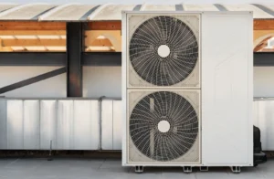 A large outdoor air conditioning unit with two fans, placed on a rooftop with ductwork in the background.