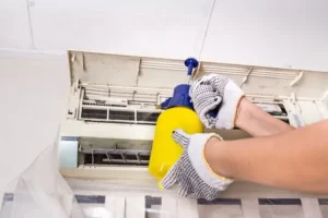 Person cleaning air conditioner filter with a yellow spray bottle, wearing gloves, emphasizing the importance of maintaining airflow for effective cooling.