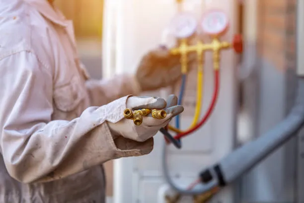 A technician, wearing a work shirt and protective gloves, holds several brass flare fittings in their left hand, with a set of manifold gauges connected to an HVAC unit visible and blurred in the background.