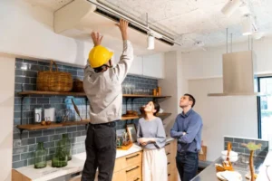 An HVAC technician in a yellow hard hat installs or maintains a ductless air conditioning unit mounted on the ceiling in a modern kitchen as a couple looks on.
