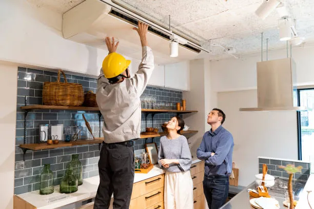 An HVAC technician in a yellow hard hat installs or maintains a ductless air conditioning unit mounted on the ceiling in a modern kitchen as a couple looks on.